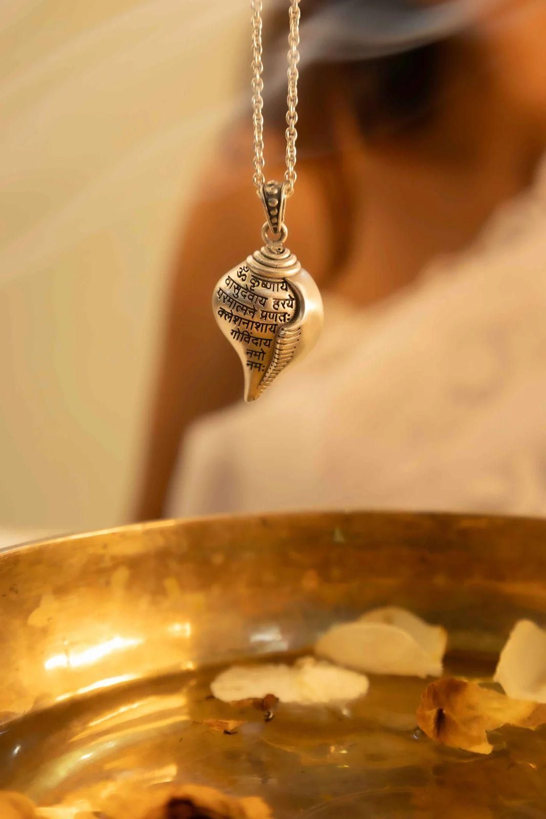 A silver-colored Shri Krishna Shankha pendant attached to a chain, displayed against a pink background.