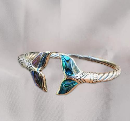 An abalone shell mermaid tail bangle bracelet displayed on a textured surface with a twisted metal band and inlayed shell details.