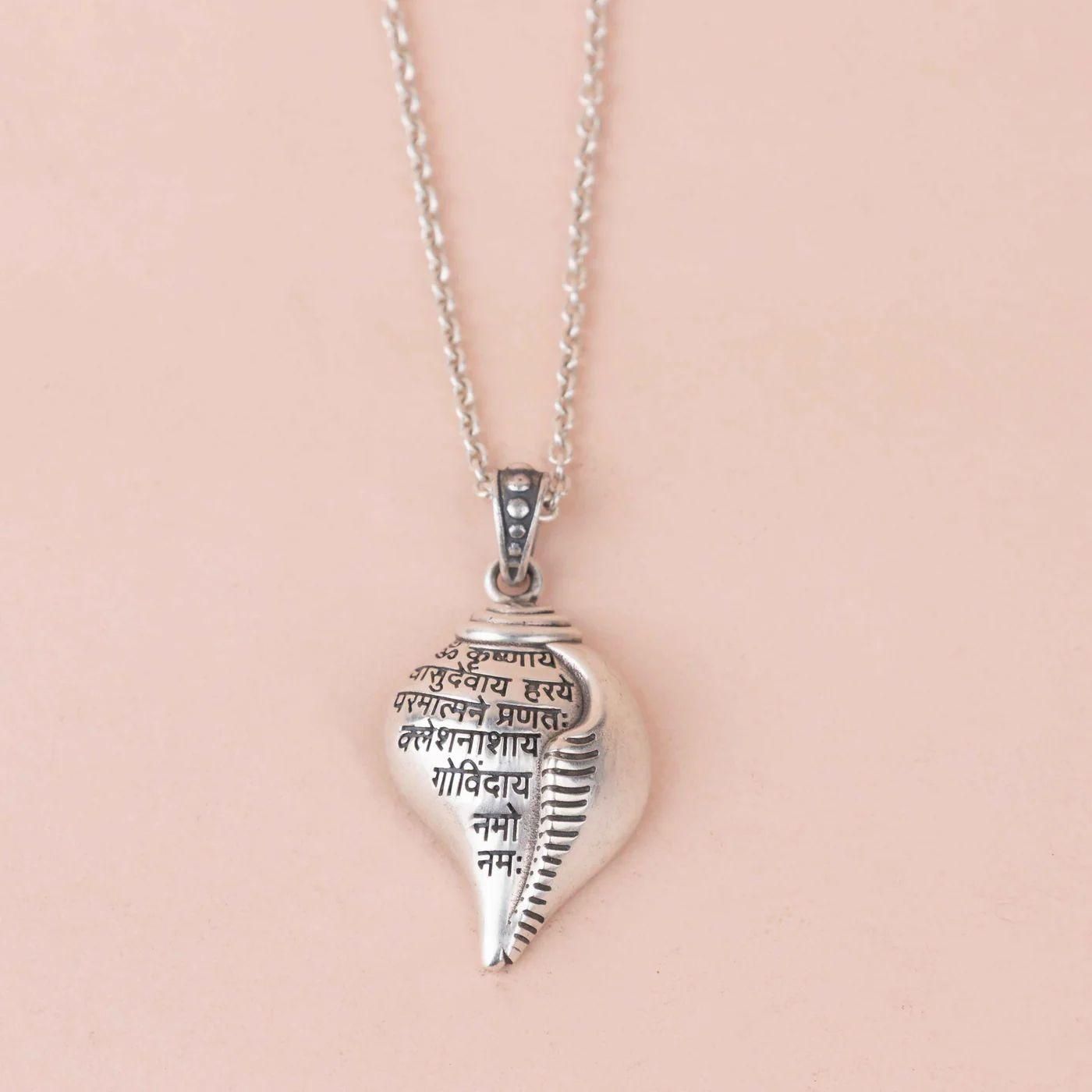 A silver-colored Shri Krishna Shankha pendant attached to a chain, displayed against a pink background.