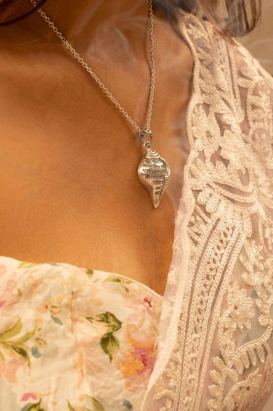 A silver-colored Shri Krishna Shankha pendant attached to a chain, displayed against a pink background.