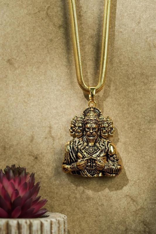 A golden-colored pendant featuring Panchamukhi Hanuman seated in a meditation pose, attached to a golden snake chain against a textured background with a pink flower and a candle.