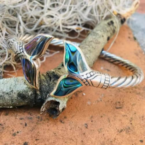 An abalone shell mermaid tail bangle bracelet displayed on a textured surface with a twisted metal band and inlayed shell details.