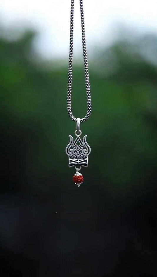 A silver oxidised Trishul Damru Rudraksha pendant chain with a small red bead on the pendant, captured against a blurred green background.