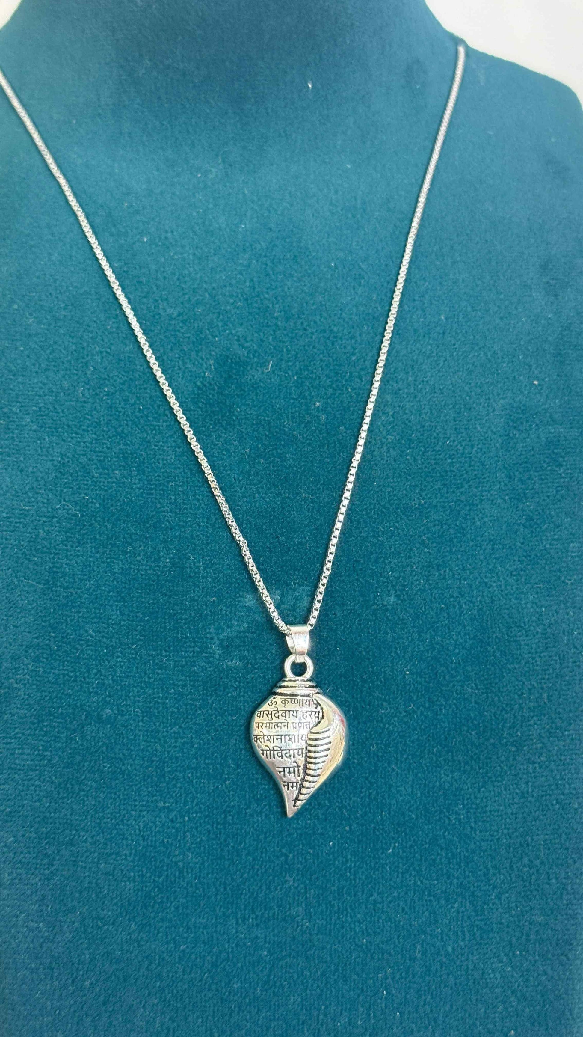 A silver-colored Shri Krishna Shankha pendant attached to a chain, displayed against a pink background.