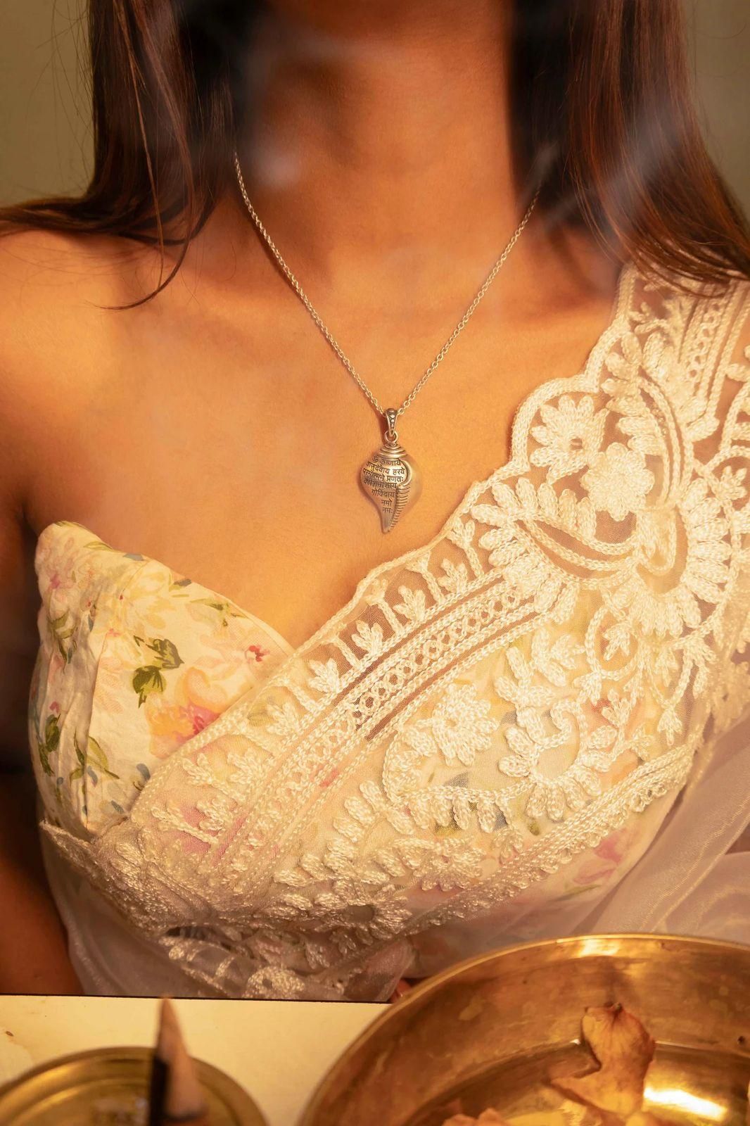 A silver-colored Shri Krishna Shankha pendant attached to a chain, displayed against a pink background.
