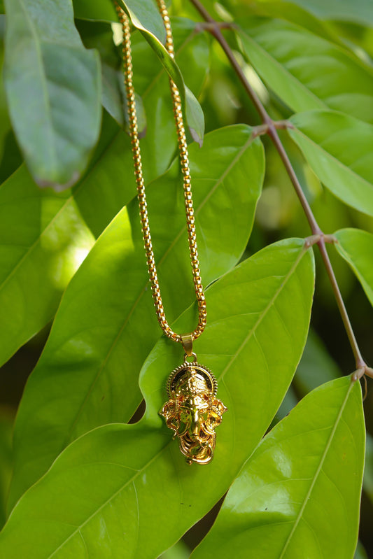A gold-colored Ganesha pendant attached to a snake chain, displayed against a backdrop of green leaves.