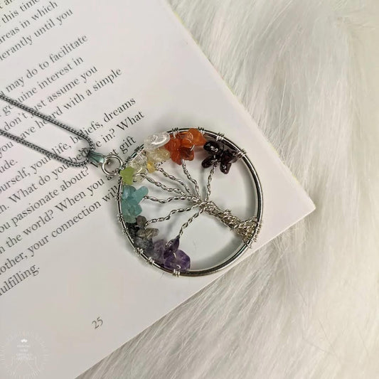 A silver-colored Tree of Life pendant with multicolored chakra stones arranged in a circle, displayed on a white surface with a book.